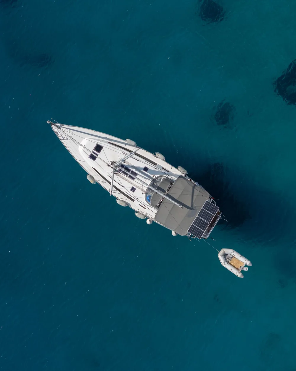 A white sailing yacht at anchor in clear turquoise water, seen from directly above, its tender astern
