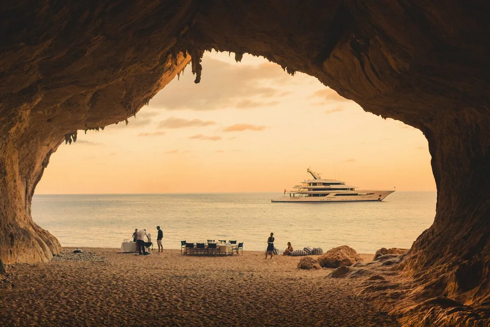 Seen from inside a sea cave at sunset, a table is set on the sand while a superyacht waits at anchor