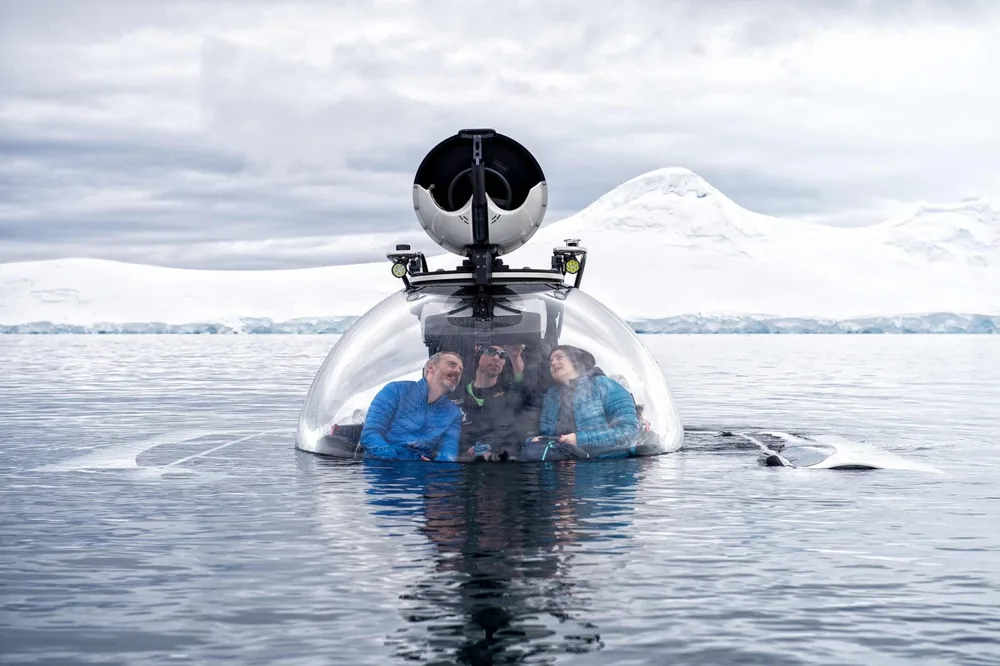 A clear-domed guest submarine surfaces among Antarctic ice with passengers inside