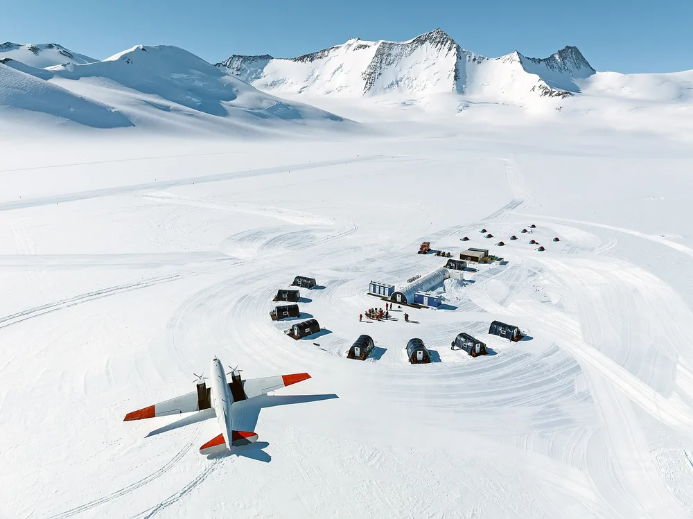 A small camp of tents on the Antarctic ice from the air, a ski plane parked beside it beneath white peaks