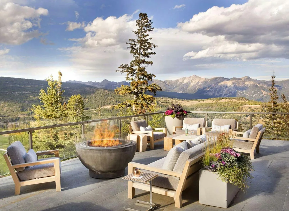 A terrace with a fire pit and lounge chairs looking out over pines to the Madison Range